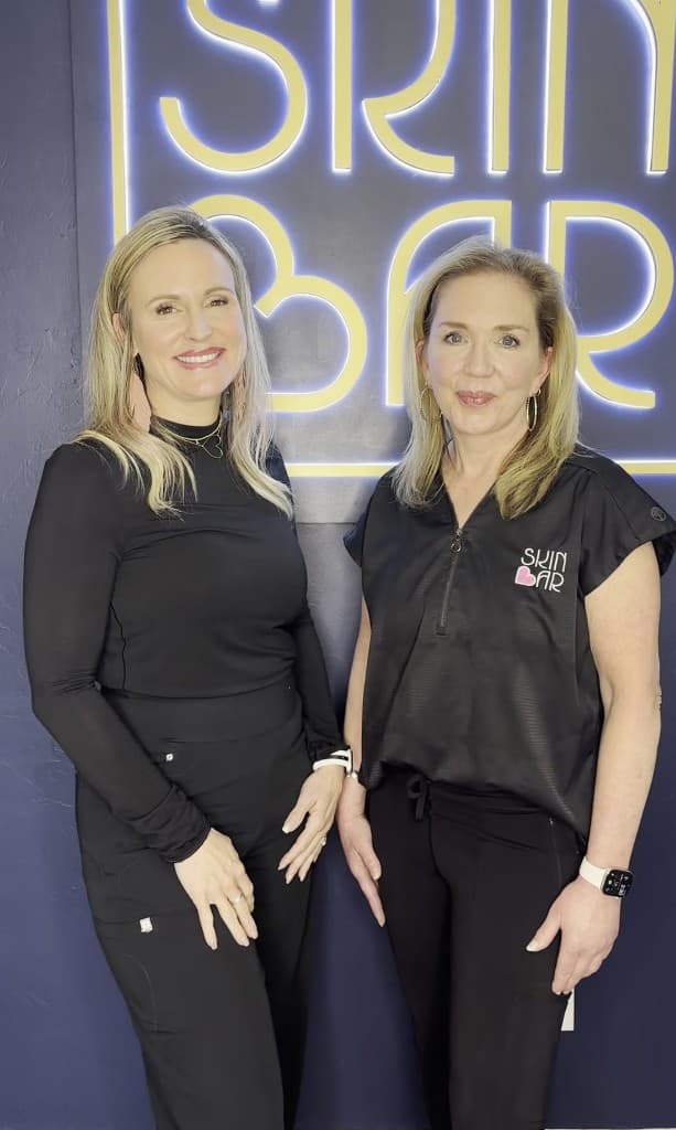 Two members of the Skin Bar OKC team in front of the neon Skin Bar sign